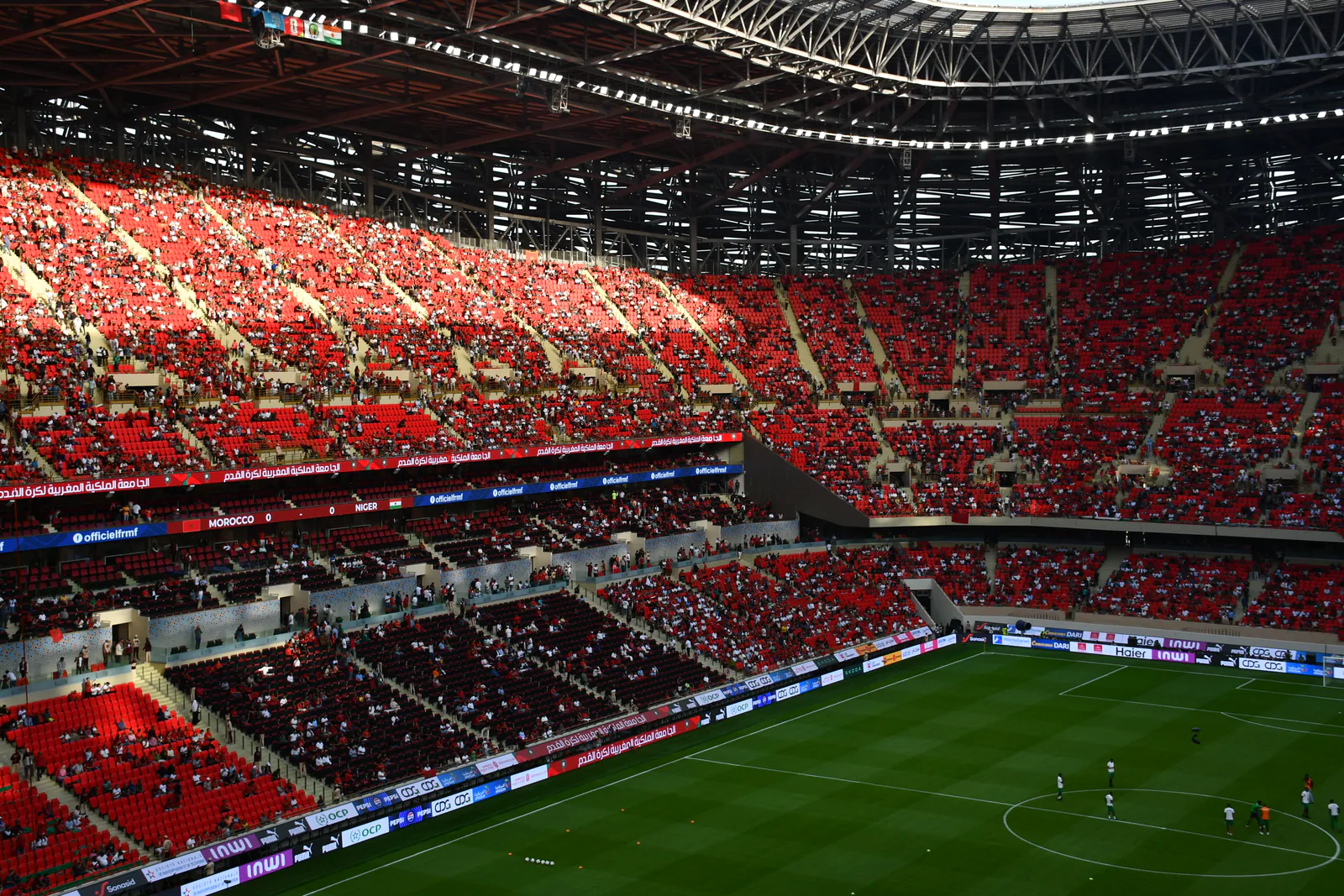 Upper-tier view across the green pitch and red seating bowl beneath the steel roof at Prince Moulay Abdellah Stadium